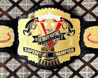May include: A gold and black UFC Superfight Champion belt. The belt features a large gold center plate with the words "THE ULTIMATE FIGHTING CHAMPIONSHIP" and "SUPERFIGHT CHAMPION". Two smaller plates on each side read "UFC WOW THE ULTIMATE FIGHTING".