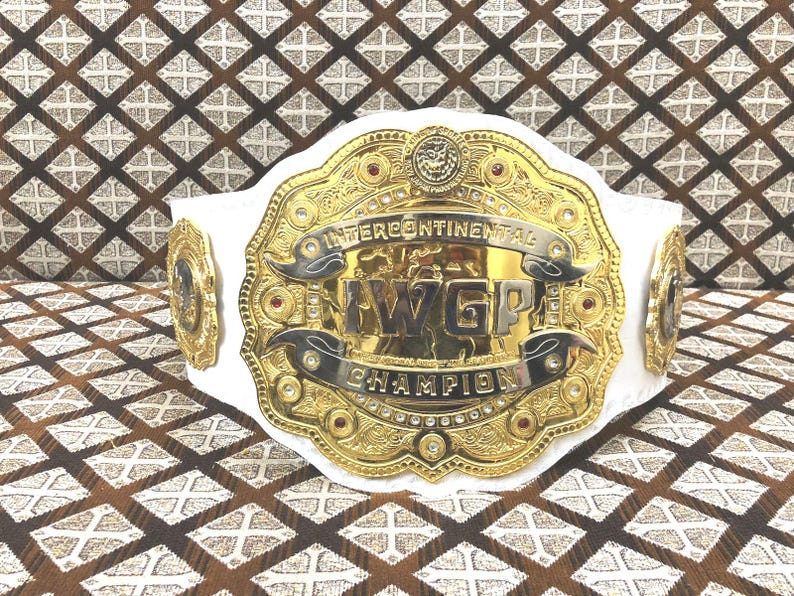 May include: A championship wrestling belt featuring a large, ornate gold-plated center plate with the text "IWGP INTERCONTINENTAL CHAMPION" engraved in bold lettering. The belt includes a white leather strap and smaller gold side plates.