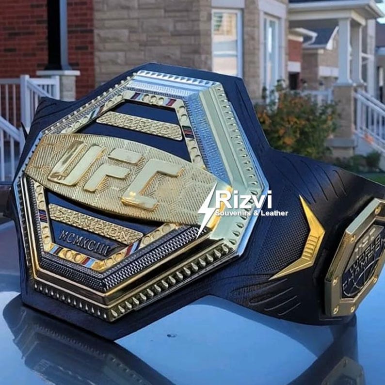 May include: A gold and black UFC championship belt with the words "CHAMPION" and "MCMXCIII" on the front. The belt is made of leather and has a hexagonal design.