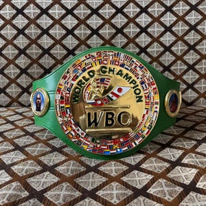 May include: A green and gold World Boxing Council (WBC) championship belt. The belt features a large central emblem with the words "WORLD CHAMPION" and "WBC". The belt is adorned with flags and portraits.