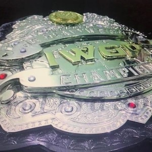 Puede incluir: Un cinturón de campeonato plateado y dorado con el texto "IWGP CHAMPION" en letras doradas. El cinturón tiene detalles intrincados, acentos rojos y medallones decorativos. El diseño general es ornamentado y llamativo.