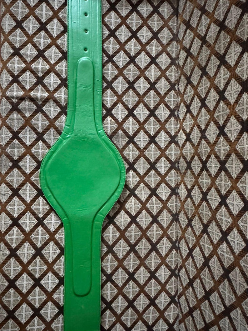 May include: A bright green leather belt with a wide, padded center section and a series of holes for adjustment. The belt is shown against a patterned brown and tan fabric background.