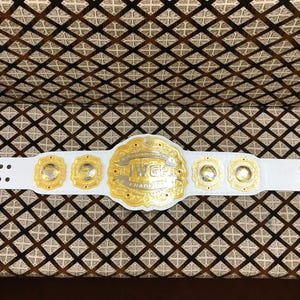 May include: A white leather championship belt featuring a large central gold plate with the text IWGP CHAMPION, flanked by four smaller circular gold plates. The belt includes gold-toned metal studs and a gold-colored buckle on the right side.