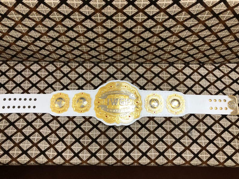 May include: A white leather championship belt featuring a large central gold plate with the text IWGP CHAMPION, flanked by four smaller circular gold plates. The belt includes gold-toned metal studs and a gold-colored buckle on the right side.