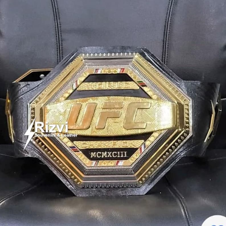 May include: A gold and black UFC championship belt with the UFC logo and the year MCMXCIII inscribed on the front. The belt is made of leather and has a octagonal design.