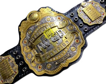Pode incluir: Um cinturão de campeonato dourado e prateado com as palavras "HEAVYWEIGHT CHAMPION" e "RIZIN" na frente. O cinturão tem uma correia de couro preta e é decorado com joias vermelhas e desenhos intrincados.