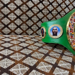 May include: A green boxing championship belt with gold accents and portraits. The belt features the words "WORLD CHAMPION" and "WBC". The background is a brown and tan patterned fabric.