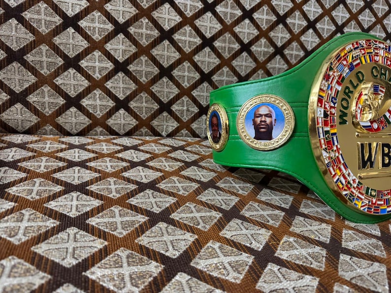 May include: A green boxing championship belt with gold accents and portraits. The belt features the words "WORLD CHAMPION" and "WBC". The background is a brown and tan patterned fabric.
