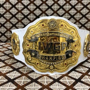 May include: A white leather championship belt featuring a large, ornate gold-plated center plate with intricate engravings and red gemstone accents. The text "INTERCONTINENTAL" and "IWGP CHAMPION" is prominently displayed in bold, raised lettering.