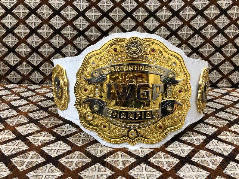May include: A white leather championship belt featuring a large, ornate gold-plated center plate with intricate engravings and red gemstone accents. The text "INTERCONTINENTAL" and "IWGP CHAMPION" is prominently displayed in bold, raised lettering.