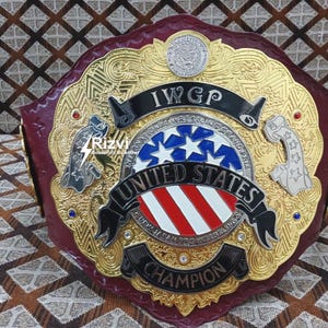 Könnte beinhalten: Ein gold- und rotfarbener Wrestling-Meisterschaftsgürtel mit der Aufschrift "IWGP United States Champion". Der Gürtel hat ein amerikanisches Flaggen-Design in Rot, Weiß und Blau.