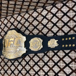 May include: A black leather UFC championship belt with gold accents and the UFC logo. The belt has multiple plates with the UFC logo and the words "Ultimate Fighting Championship".