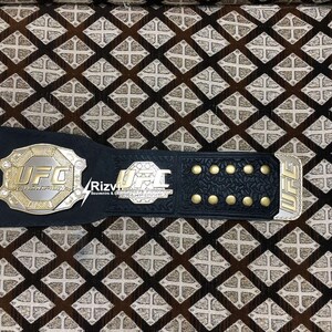 May include: A black UFC championship belt with gold accents and the word "UFC" in gold lettering. The belt has a black leather strap with gold studs.