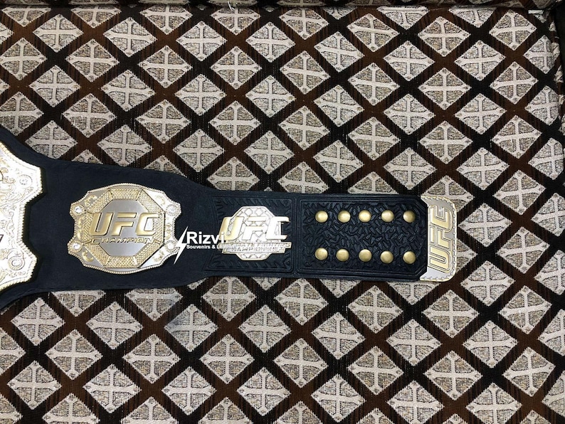 May include: A black UFC championship belt with gold accents and the word "UFC" in gold lettering. The belt has a black leather strap with gold studs.