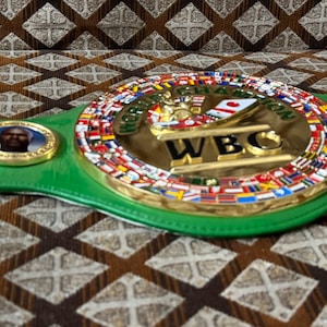 May include: A green and gold World Boxing Council (WBC) championship belt. The center plate features the WBC logo and the words "WORLD CHAMPION" and "WBC". The belt is adorned with flags and portraits.