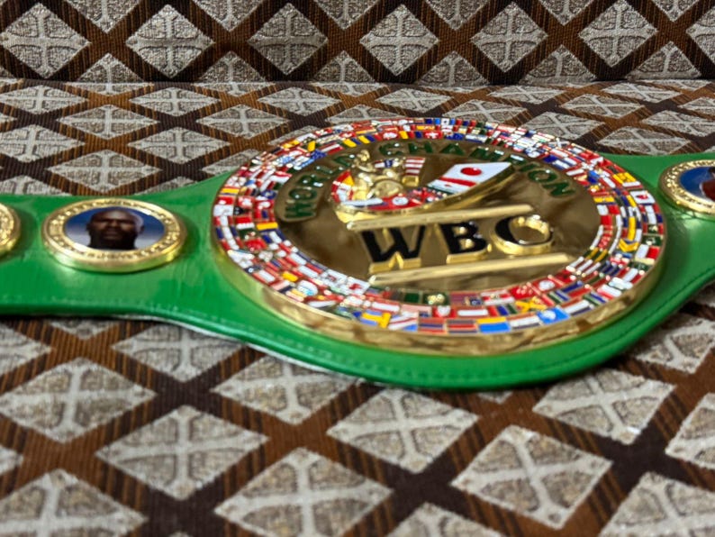 May include: A green and gold World Boxing Council (WBC) championship belt. The center plate features the WBC logo and the words "WORLD CHAMPION" and "WBC". The belt is adorned with flags and portraits.