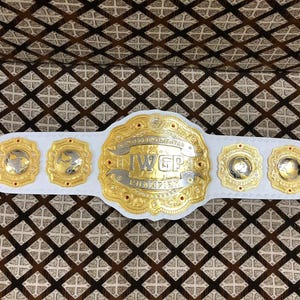 May include: A white championship belt featuring a large central gold and silver plate with the text IWGP CHAMPION. The belt includes four smaller gold and silver side plates, each displaying a globe design, all mounted on a white leather strap.