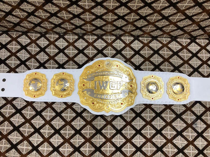 May include: A white championship belt featuring a large central gold and silver plate with the text IWGP CHAMPION. The belt includes four smaller gold and silver side plates, each displaying a globe design, all mounted on a white leather strap.