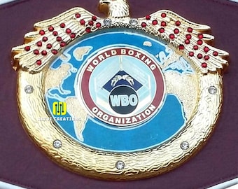 May include: A gold-colored boxing championship belt with a burgundy leather strap. The belt features a central emblem with the words "World Boxing Organization" and "WBO". An eagle with red accents sits atop the emblem, and the belt is adorned with rhinestones.