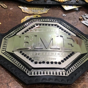 Può includere: Una cintura da campionato di wrestling professionale con una grande placca centrale argentata. La placca presenta le lettere "BMR" e la parola "MOTHERFUCKER" in grassetto. La cintura è nera con dettagli argentati e abbellimenti a forma di stella.