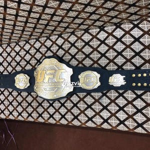 May include: A black leather UFC championship belt with gold accents and the word "UFC" in gold lettering. The belt has multiple plates with the UFC logo.