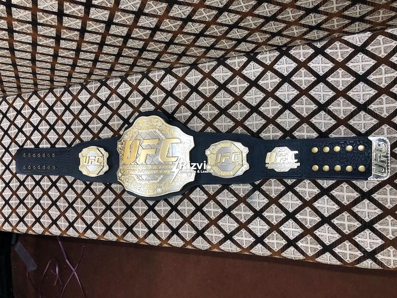 May include: A black leather UFC championship belt with gold accents and the word "UFC" in gold lettering. The belt has multiple plates with the UFC logo.