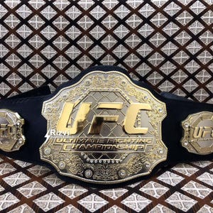 May include: A gold and black UFC championship belt with the words "Ultimate Fighting Championship" and "UFC" on the front. The belt is decorated with rhinestones and intricate designs.
