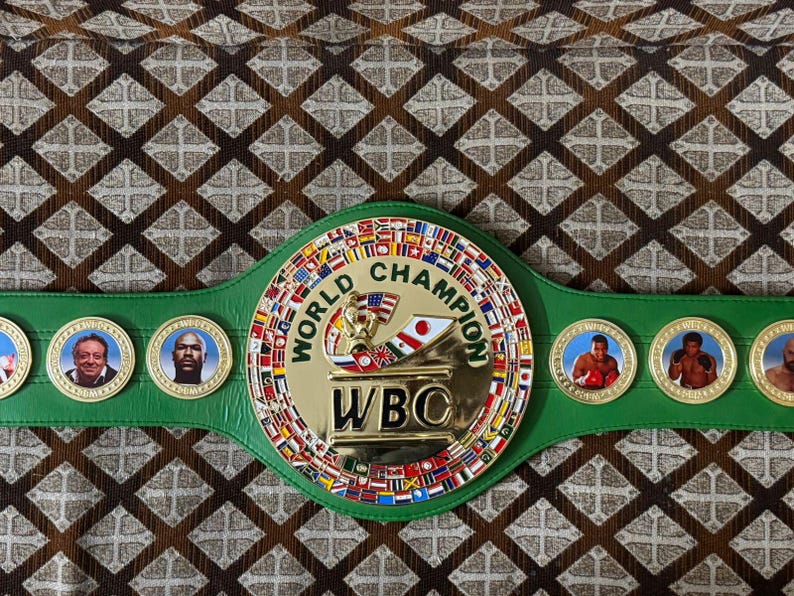 May include: A green and gold World Boxing Council (WBC) championship belt. The belt features a large central emblem with the words "World Champion" and "WBC". The belt has several medallions with portraits of boxers.