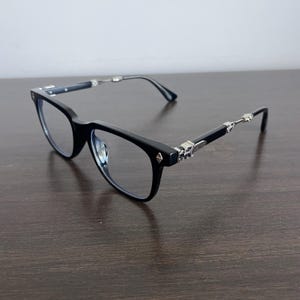 Op de afbeelding: Zwarte bril met een rechthoekig montuur en zilveren details op de pootjes. De bril heeft heldere glazen en decoratieve zilveren versieringen op de pootjes.