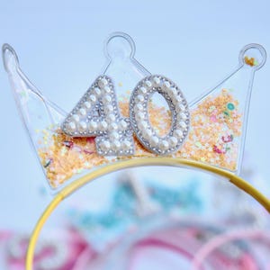 May include: A clear plastic crown headband with the number "40" decorated with white pearls and glitter. The headband is yellow and is held in a hand.