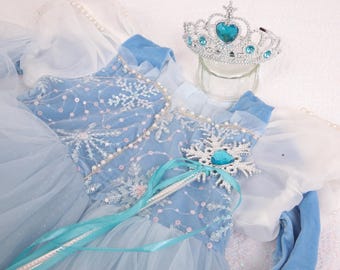 May include: A light blue princess dress with snowflake and sequin details, a silver tiara with a blue heart-shaped gem, and a matching wand. The dress has a tulle skirt and velvet bodice, perfect for dress-up and imaginative play.
