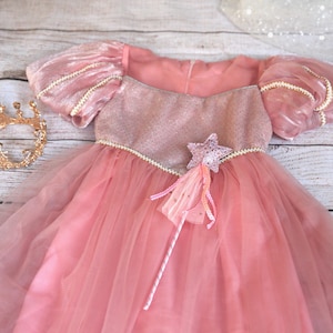 May include: A pink princess dress with a tulle skirt, puff sleeves, and a sparkly bodice. A gold crown and a star-shaped wand with ribbons are next to the dress. The dress is designed for children.