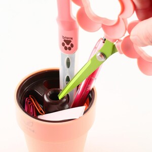 May include: A pink desk organizer shaped like a flower pot, holding pens, paperclips, and scissors. The scissors are green and pink, and the pen has the word "NIKUQ" printed on it. The organizer is peach-colored and has a dark brown interior.