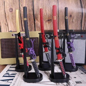 Op de afbeelding: Een verzameling miniatuur katana-zwaarden in goud, zwart en rood, elk gepresenteerd op een zwarte standaard. De zwaarden hebben decoratieve handvatten en scheden met ingewikkelde details en kleurrijke accenten. De achtergrond bevat een vintage versterker. De zwaarden zijn ongeveer 20 cm lang.