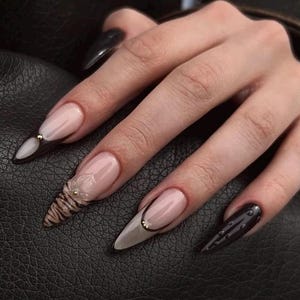 Puede incluir: Una mano con uñas acrílicas largas y puntiagudas. Las uñas presentan una variedad de diseños, incluyendo acentos negros y dorados, bases nude con puntas negras y uñas texturizadas y estampadas con adornos dorados. Un accesorio de moda.