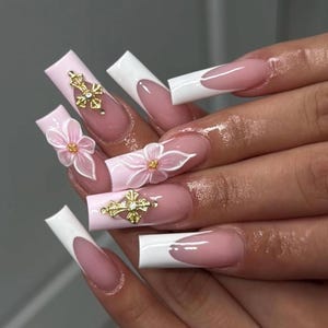 May include: A set of long, square artificial nails. The nails have a light pink base with white tips and a French manicure design. Some nails are decorated with 3D pink flowers and gold cross charms, creating a glamorous appearance.
