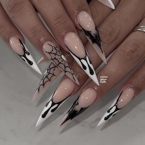 May include: Long stiletto-shaped artificial nails featuring a nude base with black drip-style French tips and a black spiderweb design on one accent nail. The text moon glow nails is printed in small white letters on the middle finger.