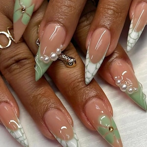 May include: Close-up of long, pointed nails with a white and green French manicure design. The nails are decorated with small, white pearls and green flowers.