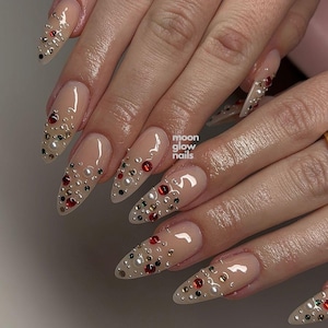 Può includere: Unghie a mandorla con una base nude lucida, decorate con un assortimento di piccoli strass rossi, verdi e neri insieme a perle bianche. Il testo moon glow nails è visibile in lettere bianche sull'unghia centrale.