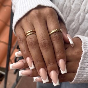 Pode incluir: Dois an&eacute;is de ouro em dedos com unhas longas de formato quadrado com um design cl&aacute;ssico de manicure francesa. Um anel &eacute; uma banda de ouro fina e lisa, enquanto o outro &eacute; uma banda de ouro tran&ccedil;ada e texturizada, ambos na mesma m&atilde;o.