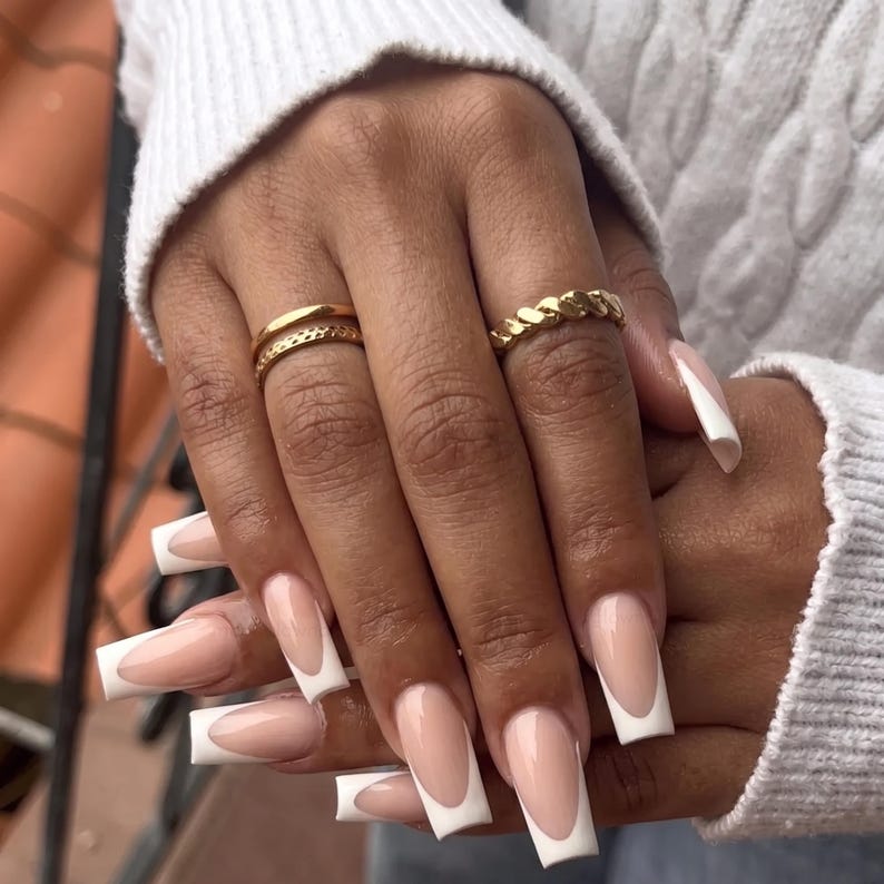 Pode incluir: Dois an&eacute;is de ouro em dedos com unhas longas de formato quadrado com um design cl&aacute;ssico de manicure francesa. Um anel &eacute; uma banda de ouro fina e lisa, enquanto o outro &eacute; uma banda de ouro tran&ccedil;ada e texturizada, ambos na mesma m&atilde;o.