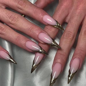 Puede incluir: Primer plano de una mano con uñas largas en forma de almendra. Las uñas están pintadas de color rosa claro y tienen una punta dorada metálica. Las uñas están bien cuidadas y tienen un acabado brillante.