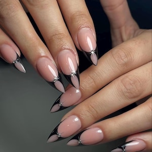 Könnte beinhalten: Lange, spitze Fingernägel mit nackter Basis und schwarzen Spitzen. Jeder Nagel hat einen kleinen, silbernen Edelstein an der Basis der Spitze. Die Nägel sind gut gepflegt und zeigen ein modernes, elegantes Design.