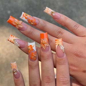 May include: A hand displaying long, square-shaped acrylic nails. The nails feature a vibrant orange and white design with floral accents and gold embellishments. Some nails have 3D flower details.