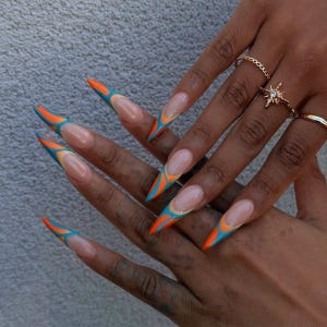 May include: Long stiletto-shaped nails featuring a French manicure style with a nude base and vibrant orange and blue abstract flame-like patterns at the tips. Three gold-toned rings are worn on the fingers, including one with a starburst design.