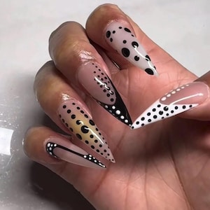May include: Long, pointed stiletto-shaped artificial nails featuring a nude base color. The nail art designs include black and white polka dots, intricate black and white line work, and French-style tips with varying dot patterns across each nail.