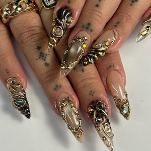May include: Close-up of long, pointed fingernails with intricate gold and jewel embellishments. The nails feature a nude base with ornate gold designs, including swirls, frames, and embedded gemstones. Rings with gold designs are also visible.
