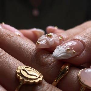May include: Nails featuring a glossy nude base with raised white 3D floral relief designs and small gold metallic beads. The manicure is complemented by three gold rings, including one with a textured oval face and another with a smooth pink stone.