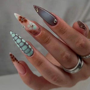 Puede incluir: Uñas largas y puntiagudas con diseños intrincados. Una uña presenta un patrón de escamas turquesa y negro, mientras que otra tiene un diseño floral con pequeños adornos plateados. Una tercera uña es de color marrón oscuro con detalles dorados.