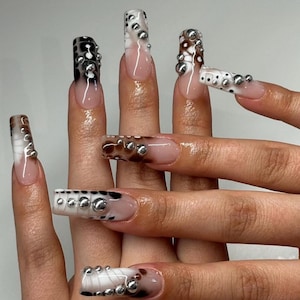May include: Long square-shaped artificial nails featuring a French manicure base with a gradient of black, brown, and white marbled patterns. The nails are decorated with small, shiny silver metallic spheres arranged in various patterns across the surface.
